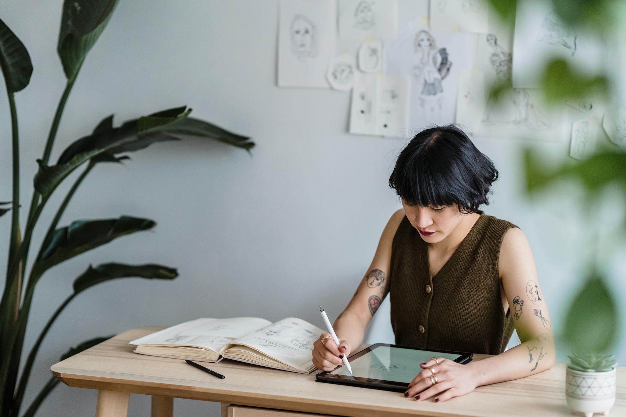 Concentrated Asian female artist sitting at table with graphics tablet and drawing sketches in creative workspace